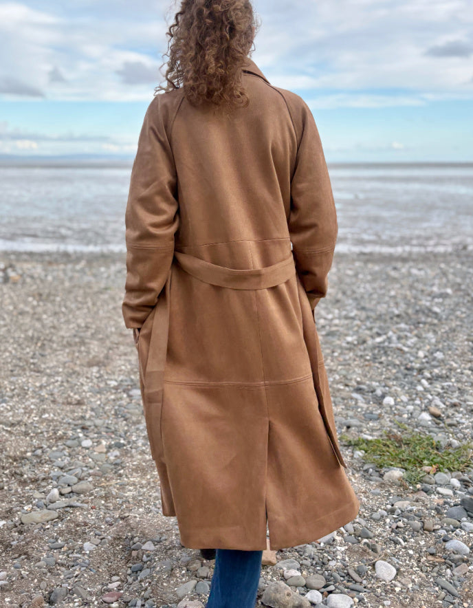 Person wearing a camel faux suede trench coat standing on a beach with a cloudy sky.