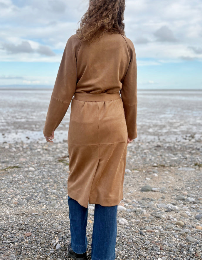 Person wearing a camel faux suede trench coat standing on a beach with a cloudy sky.