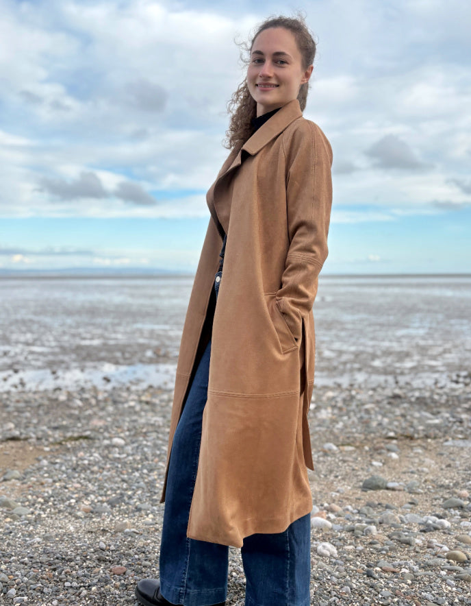 Person wearing a camel faux suede trench coat standing on a beach with a cloudy sky.