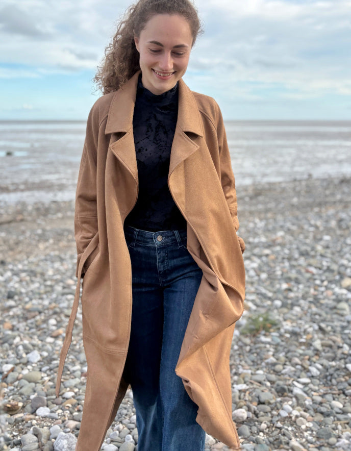 Person wearing a camel faux suede trench coat standing on a beach with a cloudy sky.