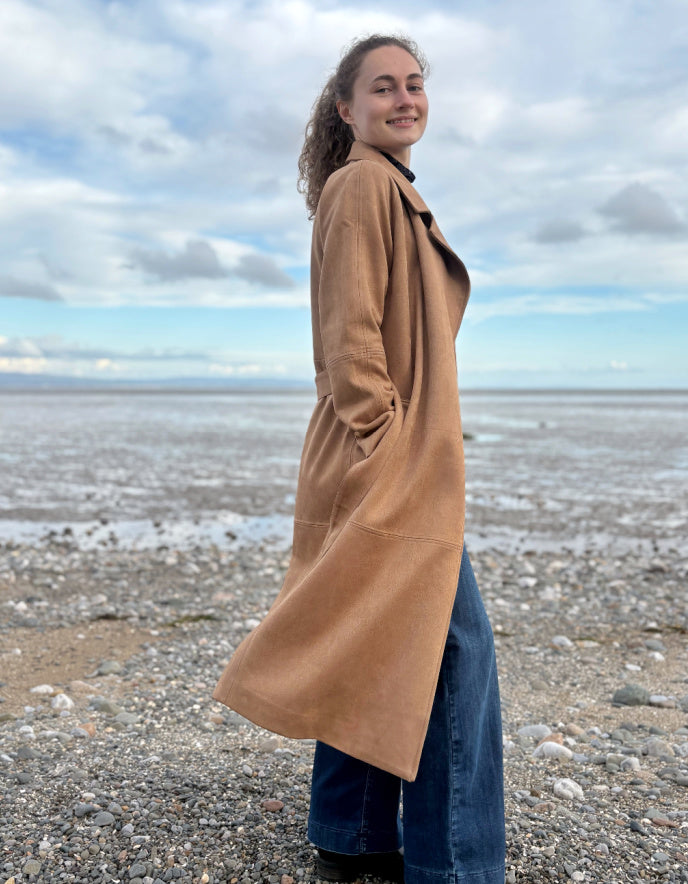 Person wearing a camel faux suede trench coat standing on a beach with a cloudy sky.
