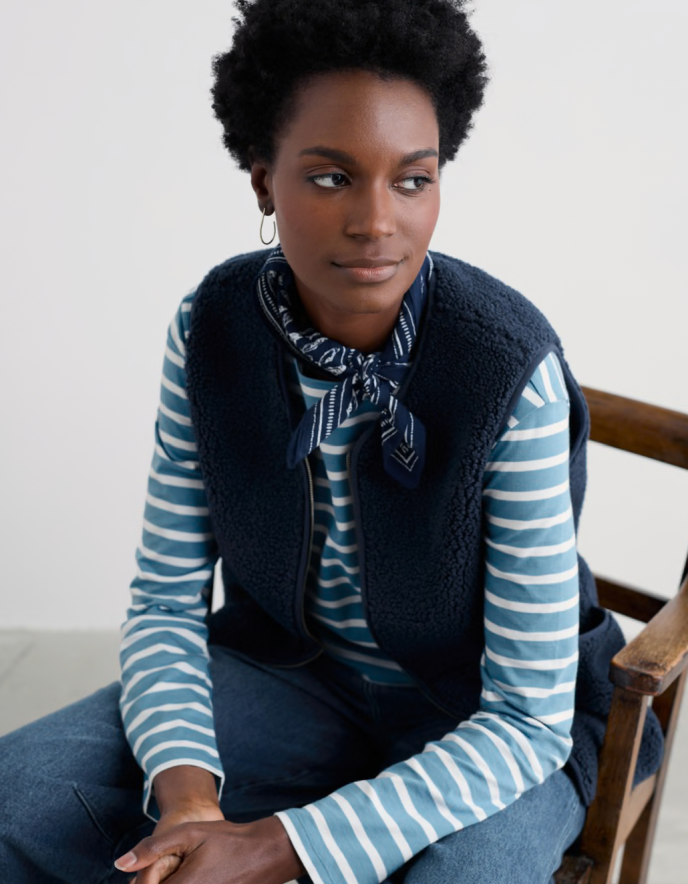 Woman wearing a blue and white striped sweater with a navy vest and scarf, sitting on a wooden chair.