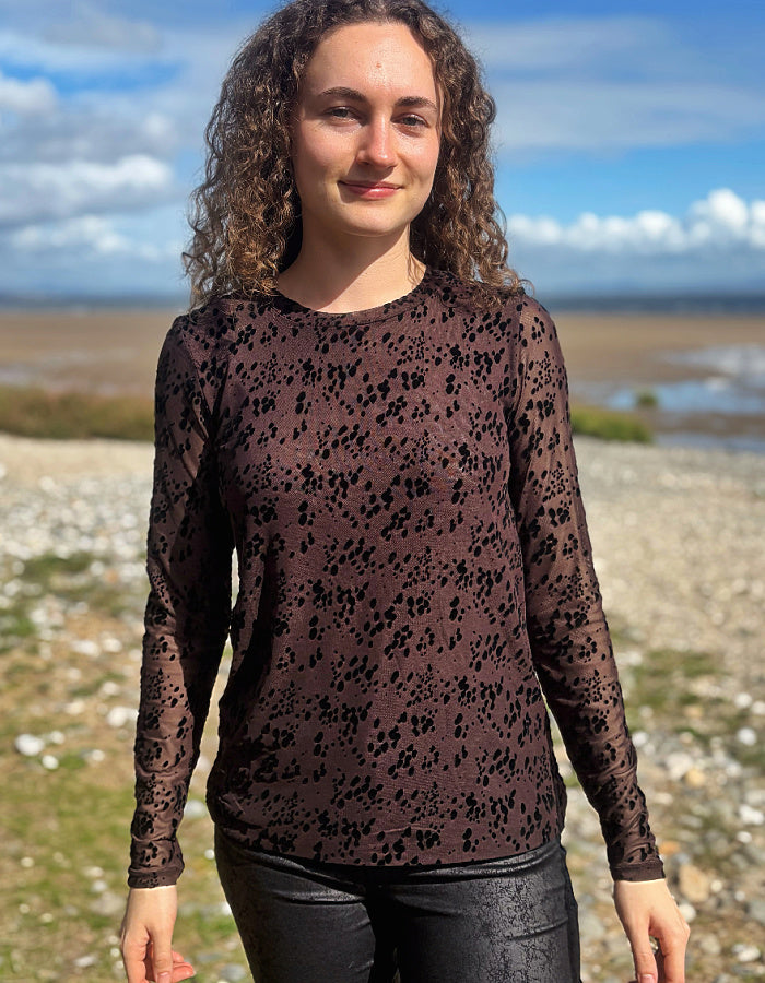 A woman with curly brown hair wearing a dark brown long-sleeve top with a black abstract pattern, standing outdoors on a rocky beach with water and a cloudy sky in the background.