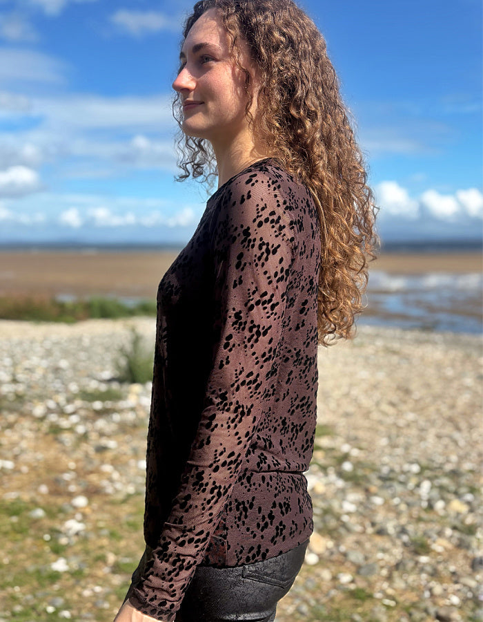 A woman with long curly brown hair stands in profile outdoors on a pebble beach, wearing a sheer long-sleeve top with a black leopard print over a dark brown base.