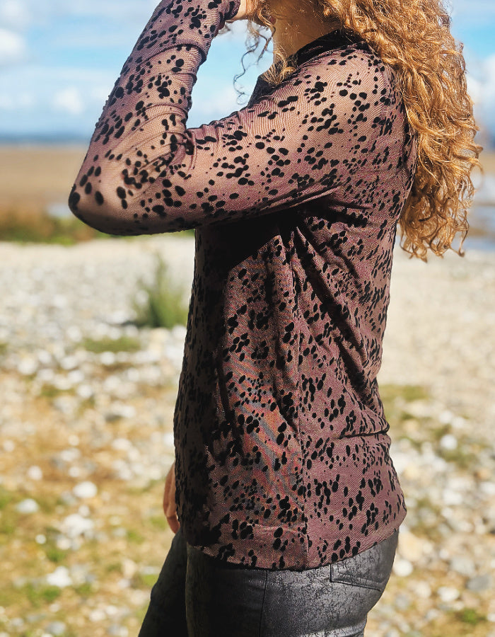 A woman wearing a sheer long-sleeve top with a black leopard print over a dusty rose base, photographed outdoors on a beach or pebble shore.