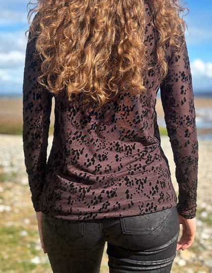 A woman viewed from behind wearing a long-sleeve sheer mesh top with a black leopard print over a dusty mauve-brown base, paired with black jeans, set against an outdoor beach background.