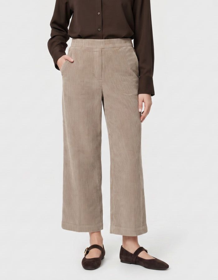 A person wearing high-waisted, wide-leg cropped trousers in a light taupe corduroy fabric, paired with a dark brown button-up shirt and dark brown flat shoes, against a plain white studio background.