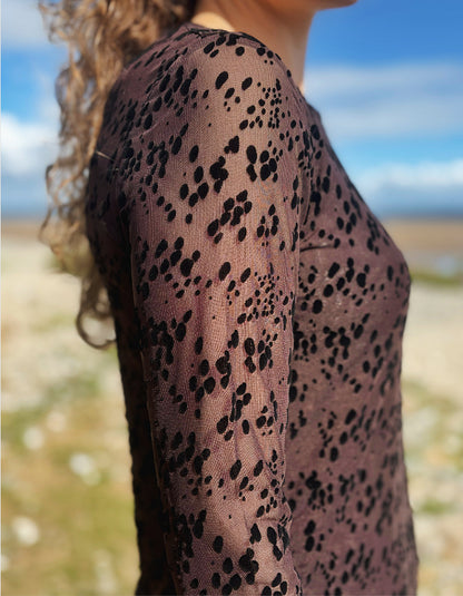 A close-up side view of a person wearing a sheer long-sleeve top with a black spotted pattern over a muted mauve-brown base, photographed outdoors against a blurred beach and sky background.