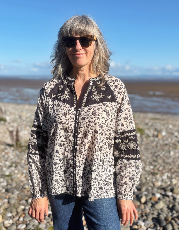 A woman wearing a navy and white floral patterned long-sleeve cotton blouse stands on a rocky beach with the ocean and blue sky in the background.
