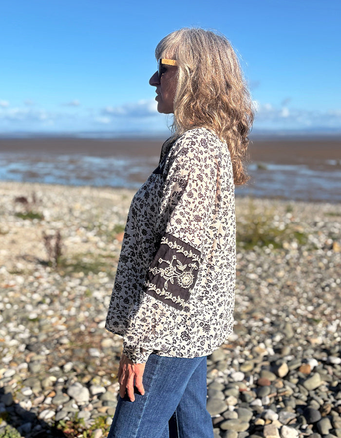 A woman wearing a navy and white floral patterned long-sleeve cotton blouse stands on a rocky beach with the ocean and blue sky in the background.