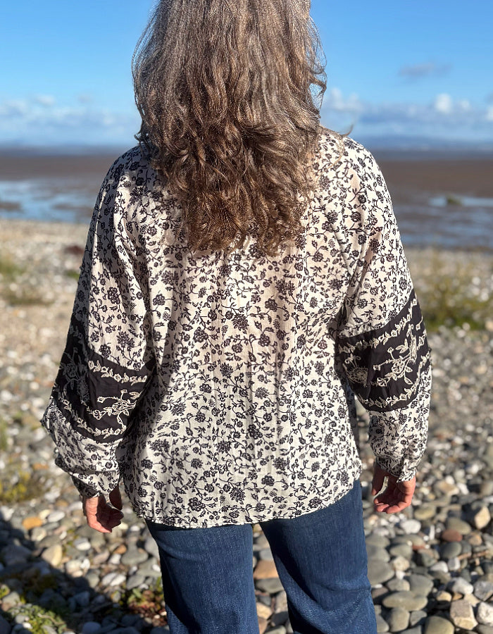 A woman viewed from behind wearing a navy and white floral blouse with patterned sleeve cuffs, standing on a pebble beach with water and sky in the background.