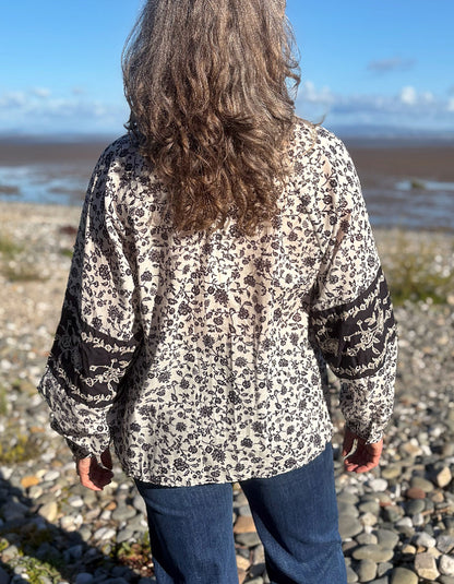 A woman viewed from behind wearing a navy and white floral blouse with patterned sleeve cuffs, standing on a pebble beach with water and sky in the background.