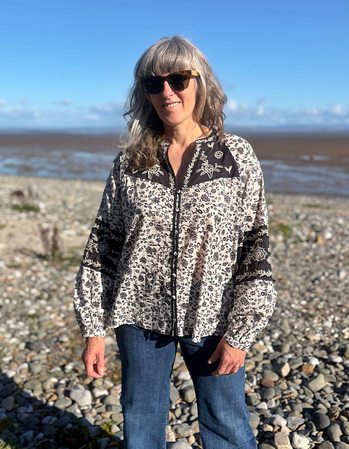 A woman wearing a navy and white floral patterned long-sleeve cotton blouse stands on a rocky beach with the ocean and blue sky in the background.