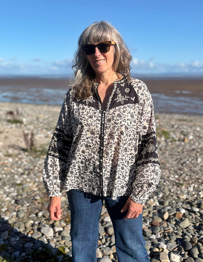 A woman wearing a navy and white floral patterned long-sleeve cotton blouse stands on a rocky beach with the ocean and blue sky in the background.