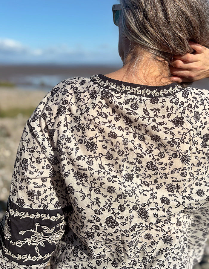 A woman wearing a navy and white floral patterned long-sleeve cotton blouse stands on a rocky beach with the ocean and blue sky in the background.