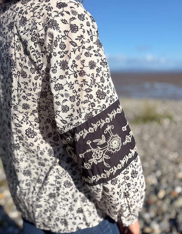 A close-up of a person wearing a cream-coloured long-sleeve blouse with a navy floral print and a contrasting dark brown sleeve panel featuring white embroidery, set against a blurred outdoor beach background.
