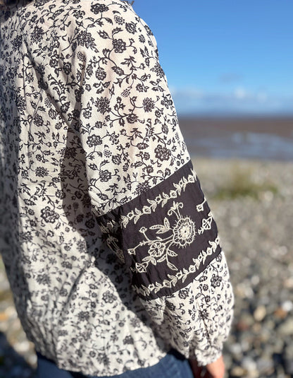 A close-up of a person wearing a cream-coloured long-sleeve blouse with a navy floral print and a contrasting dark brown sleeve panel featuring white embroidery, set against a blurred outdoor beach background.