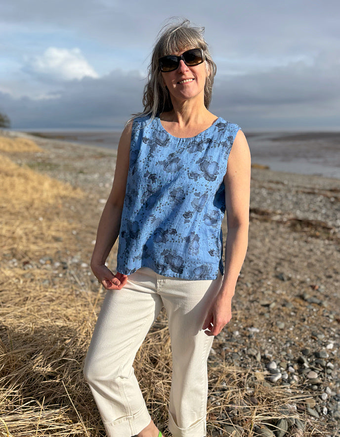 Woman wearing a blue sleeveless top and sunglasses standing in a field with a scenic background.