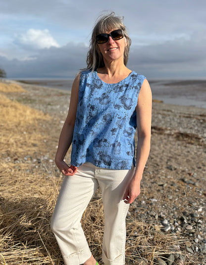 Woman wearing a blue sleeveless top and sunglasses standing in a field with a scenic background.