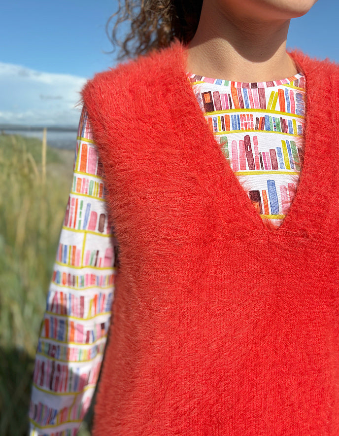 Person wearing an orange fluffy knitted vest over a colourful patterned shirt on a beach.