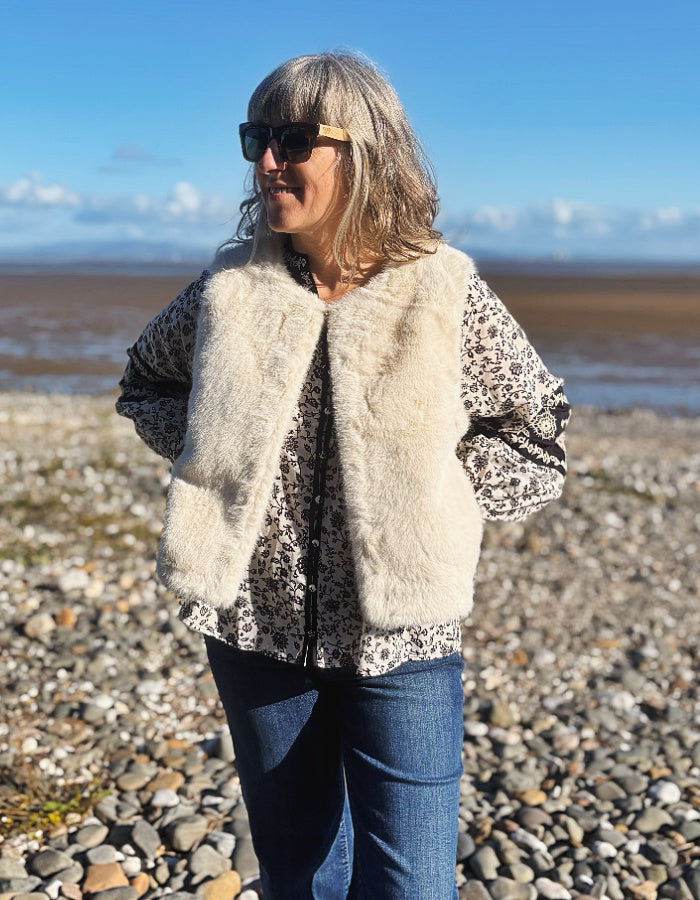 A woman wearing a cream faux fur vest over a paisley print top and blue jeans, standing on a pebble beach under a blue sky.