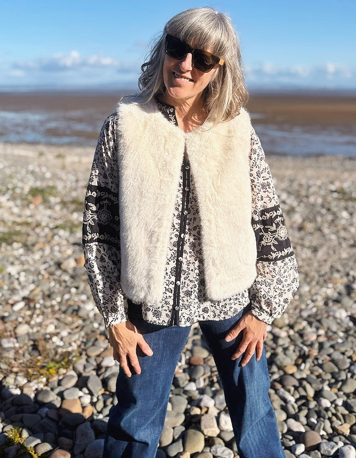 A woman wearing a cream faux-fur vest over a black and white patterned long-sleeve top and blue jeans, standing on a rocky beach with water and sky in the background.