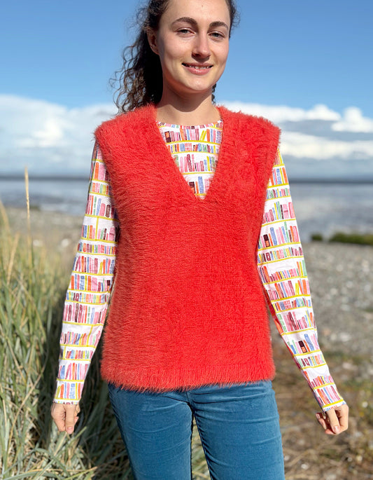 Person wearing an orange fluffy knitted vest over a colourful patterned shirt on a beach.