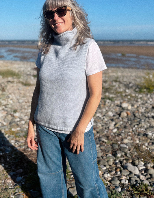 A woman wearing a light gray sleeveless ribbed turtleneck sweater vest over a white t-shirt and blue jeans, standing on a rocky beach with water and sky in the background.