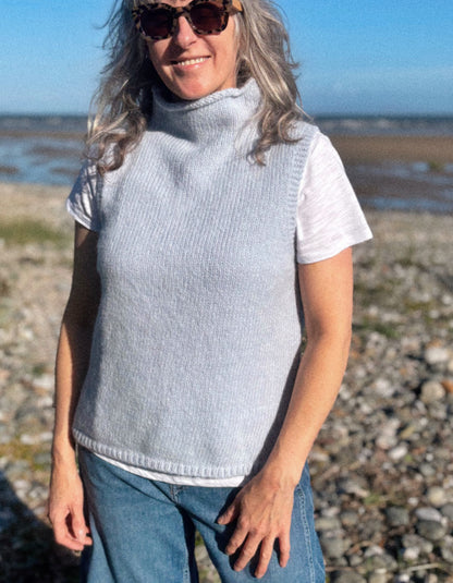 A woman wearing a light gray ribbed knit turtleneck vest over a white t-shirt and blue jeans, standing outdoors on a rocky beach with water and sky in the background.