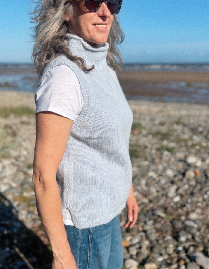 A woman wearing a light gray ribbed knit turtleneck vest over a white t-shirt and blue jeans, standing outdoors on a rocky beach with water and sky in the background.