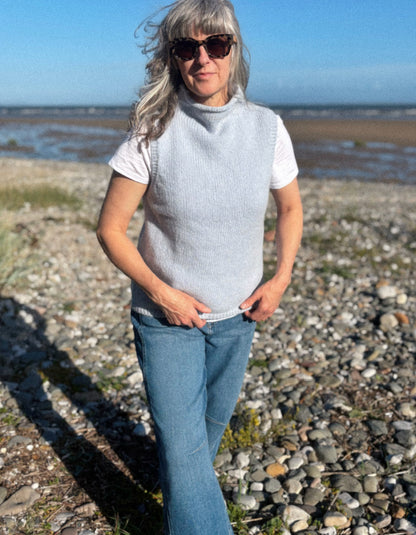A woman standing on a rocky beach wearing a light gray ribbed knit mock-neck sweater vest over a white t-shirt and blue jeans.