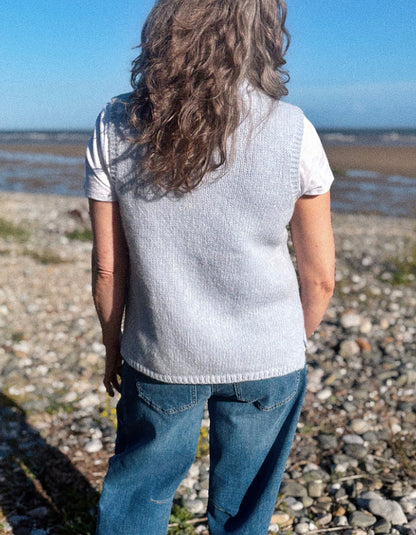 A woman viewed from behind wearing a light gray knit sleeveless vest over a white t-shirt and blue jeans, standing on a pebble beach with water and sky in the background.