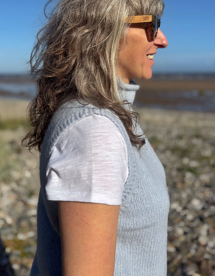 A woman in profile wearing a light blue ribbed knit sleeveless top over a white t-shirt, standing outdoors on a rocky beach with water and sky in the background.
