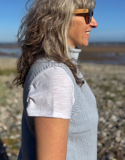 A woman in profile wearing a light blue ribbed knit sleeveless top over a white t-shirt, standing outdoors on a rocky beach with water and sky in the background.