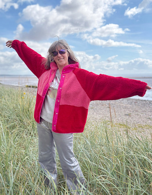 Person wearing a red and pink fleece jacket with silver wide leg jeans standing in grass with a blue sky and clouds in the background