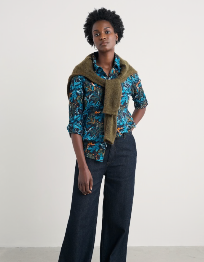 Woman wearing a floral blouse, dark pants, and a green scarf against a white background