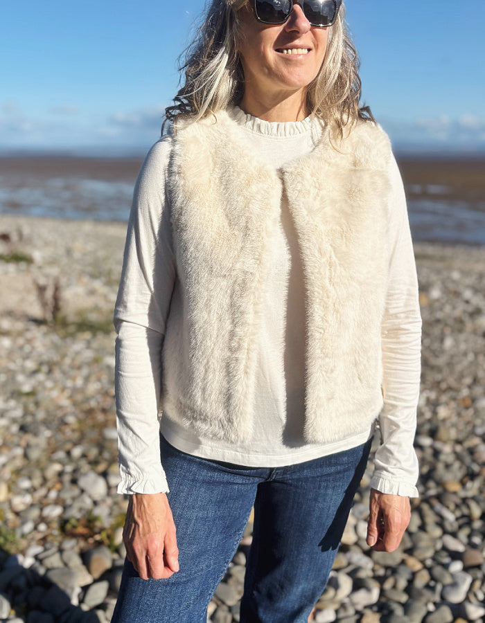 A woman wearing a cream faux fur vest over a matching cream long-sleeve top and blue jeans, standing on a rocky beach with water and sky in the background.