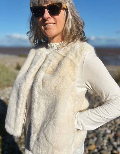 A woman outdoors wearing a cream-colored faux fur vest over a white long-sleeve top, with dark sunglasses and a blurred natural background.