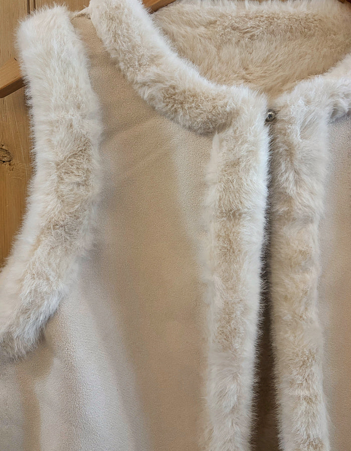 Close-up of a sleeveless cream-colored vest with thick, fluffy faux-fur trim around the collar, armholes, and front placket, shown hanging on a wooden hanger against a light wood background.
