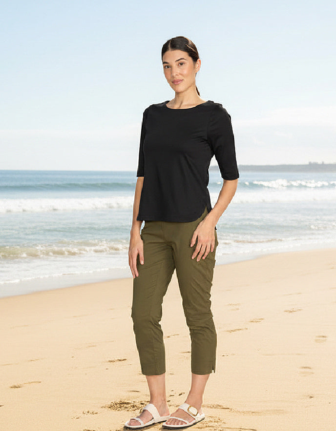 Woman standing on a beach wearing a black top and green pants.