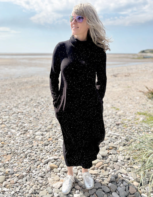 Person wearing a black dress with white speckled galactic print standing on a pebbly beach with a blue sky.