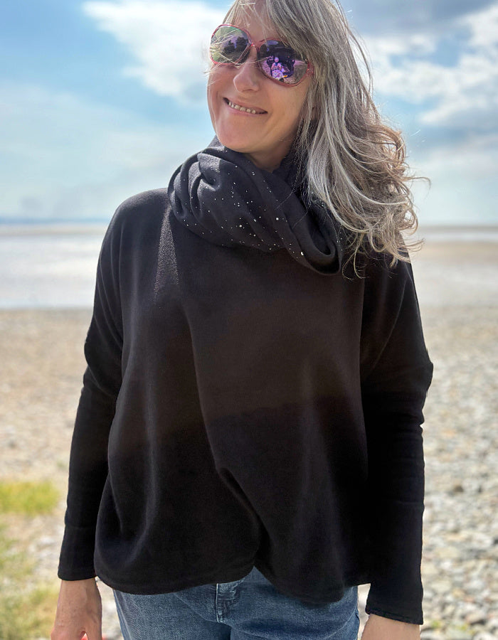 Person wearing a black sweater and snood and sunglasses on a beach