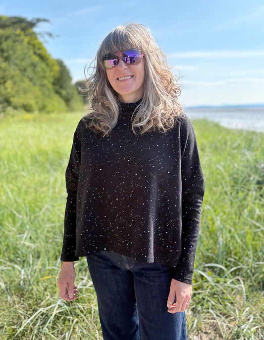 Person wearing a black speckled sweater standing in a grassy field with a scenic background.