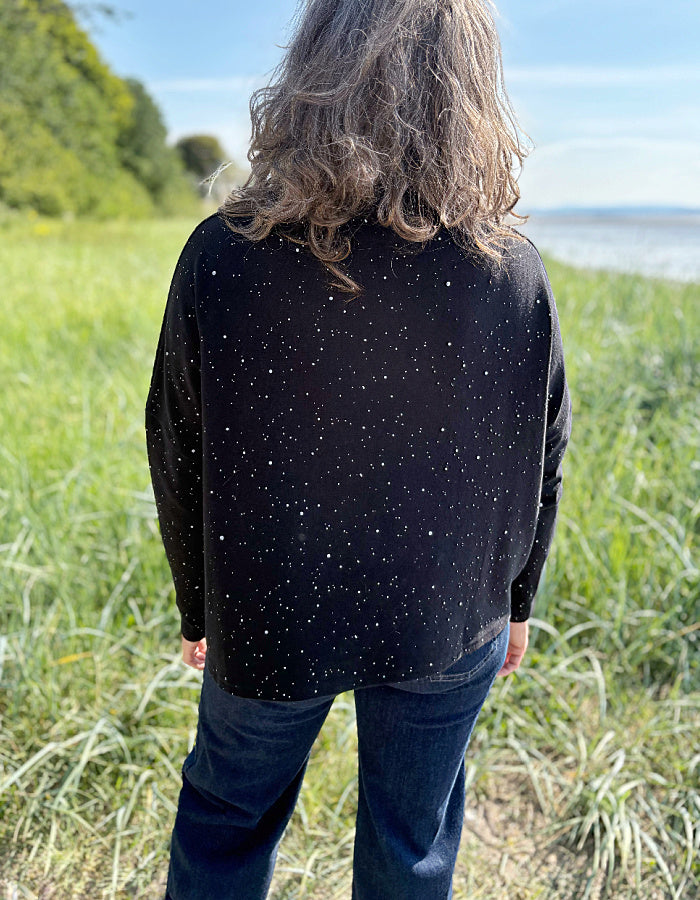 Person wearing a black speckled sweater standing in a grassy field with a body of water in the background.