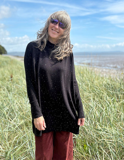 Person standing in a grassy field with a scenic background wearing a black cosy turtle neck tunic and copper brown cord trousers