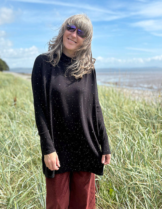 Person standing in a grassy field with a scenic background wearing a black cosy turtle neck tunic and copper brown cord trousers