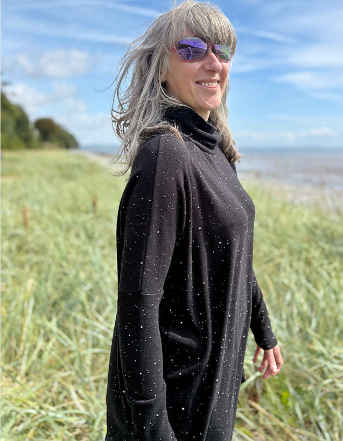 Person standing in a grassy field with a scenic background wearing a black cosy turtle neck tunic and copper brown cord trousers