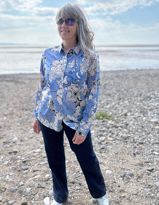 Person wearing a blue patterned shirt on a pebbly beach with water and sky in the background