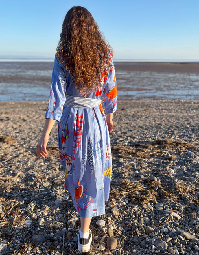 Woman standing on a beach wearing a colourful dress with a pattern of coffee cups and saucers.