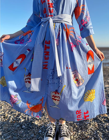 Person wearing a blue dress with food-themed graphics on a pebbly beach.